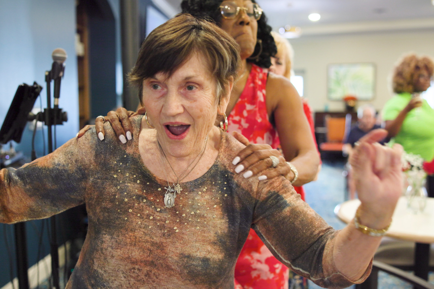 A resident in a shimmering colorful top laughs joyfully as the entertainer rests her hands on her shoulders during a lively conga line, reflecting the fun senior living with social activities at senior living in Cooper City, FL.