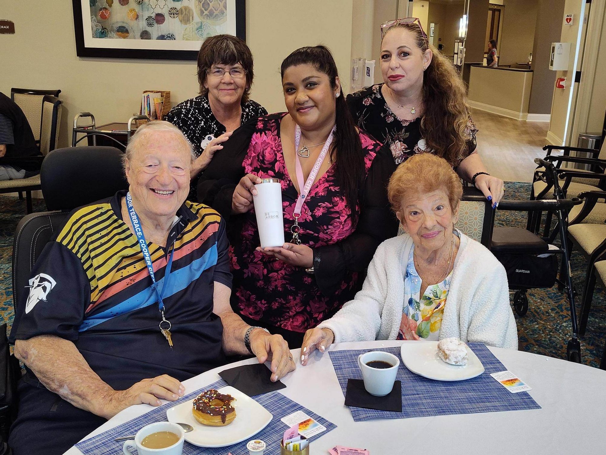 Residents enjoy coffee and dessert at a cozy table while smiling staff members gather around them, reflecting the warm atmosphere of senior living in Cooper City, FL, where senior living with social activities creates meaningful connections daily.