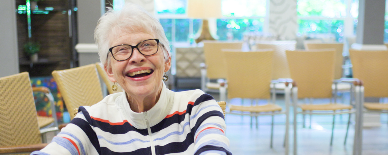 A smiling senior woman with white hair shares a joyful moment in a bright, welcoming common area, reflecting the warmth of senior living and an active senior lifestyle at The Arbor Company.