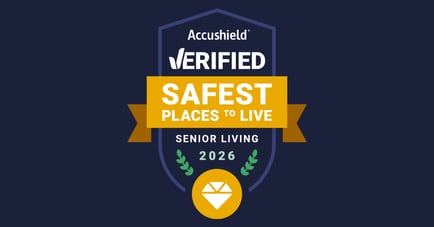 A navy blue shield-shaped badge from Accushield. The top reads 'Accushield® VERIFIED' in white text. A gold ribbon banner across the center displays 'SAFEST PLACES TO LIVE' in bold white text. Below the banner, white text reads 'SENIOR LIVING' and green laurel branches flank the year '2026' in green. At the bottom of the shield is a gold circle containing a white diamond gem icon.