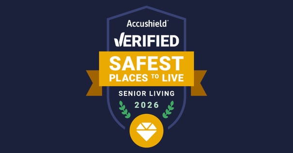 A navy blue shield-shaped badge from Accushield. The top reads 'Accushield® VERIFIED' in white text. A gold ribbon banner across the center displays 'SAFEST PLACES TO LIVE' in bold white text. Below the banner, white text reads 'SENIOR LIVING' and green laurel branches flank the year '2026' in green. At the bottom of the shield is a gold circle containing a white diamond gem icon.