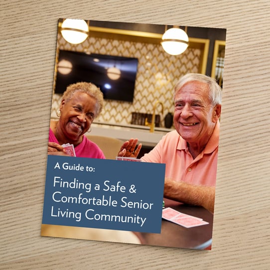 Cover of a guide titled 'A Guide to: Finding a Safe &amp; Comfortable Senior Living Community,' featuring a smiling older woman and man playing cards together in a bright common area.