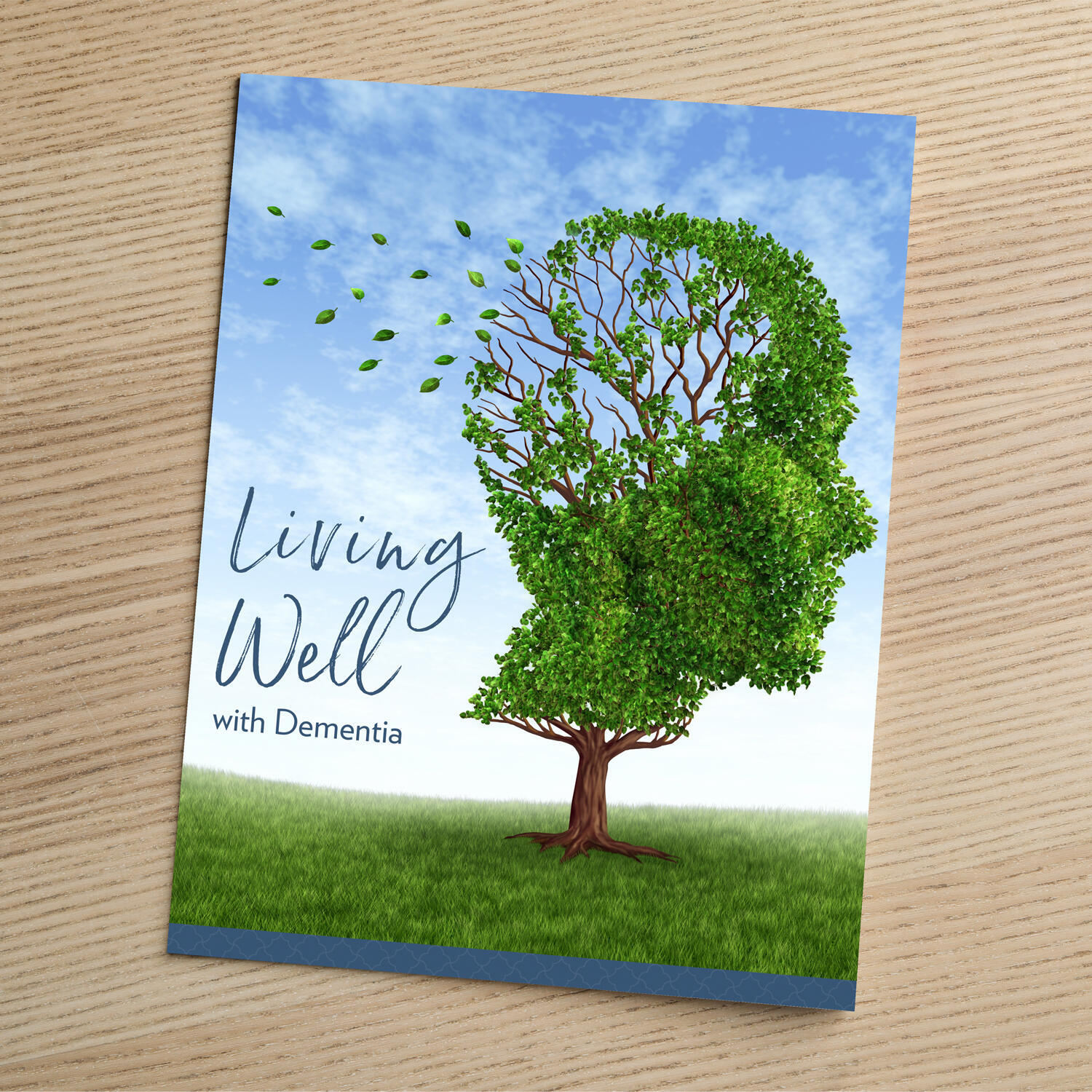 A guide titled "Living Well with Dementia" resting on a wooden surface, featuring an illustrated tree whose leafy canopy is shaped like a human head in profile, with leaves blowing away from one side to symbolize memory loss.