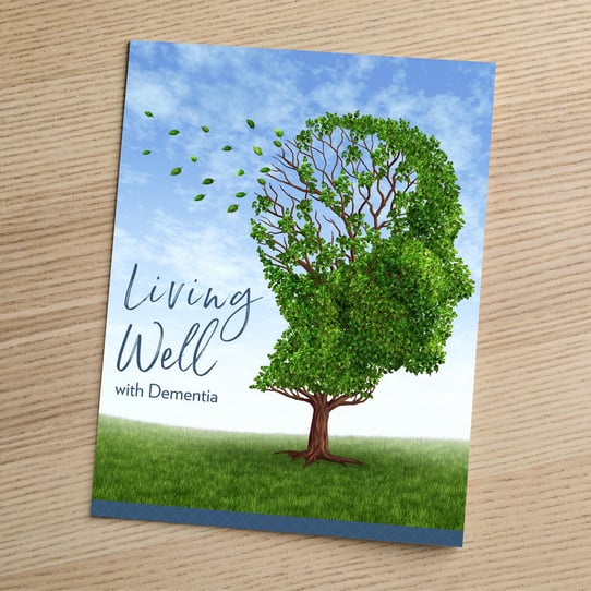 A booklet titled "Living Well with Dementia" resting on a wooden surface, featuring an illustrated tree whose leafy canopy is shaped like a human head in profile, with leaves blowing away from one side to symbolize memory loss.