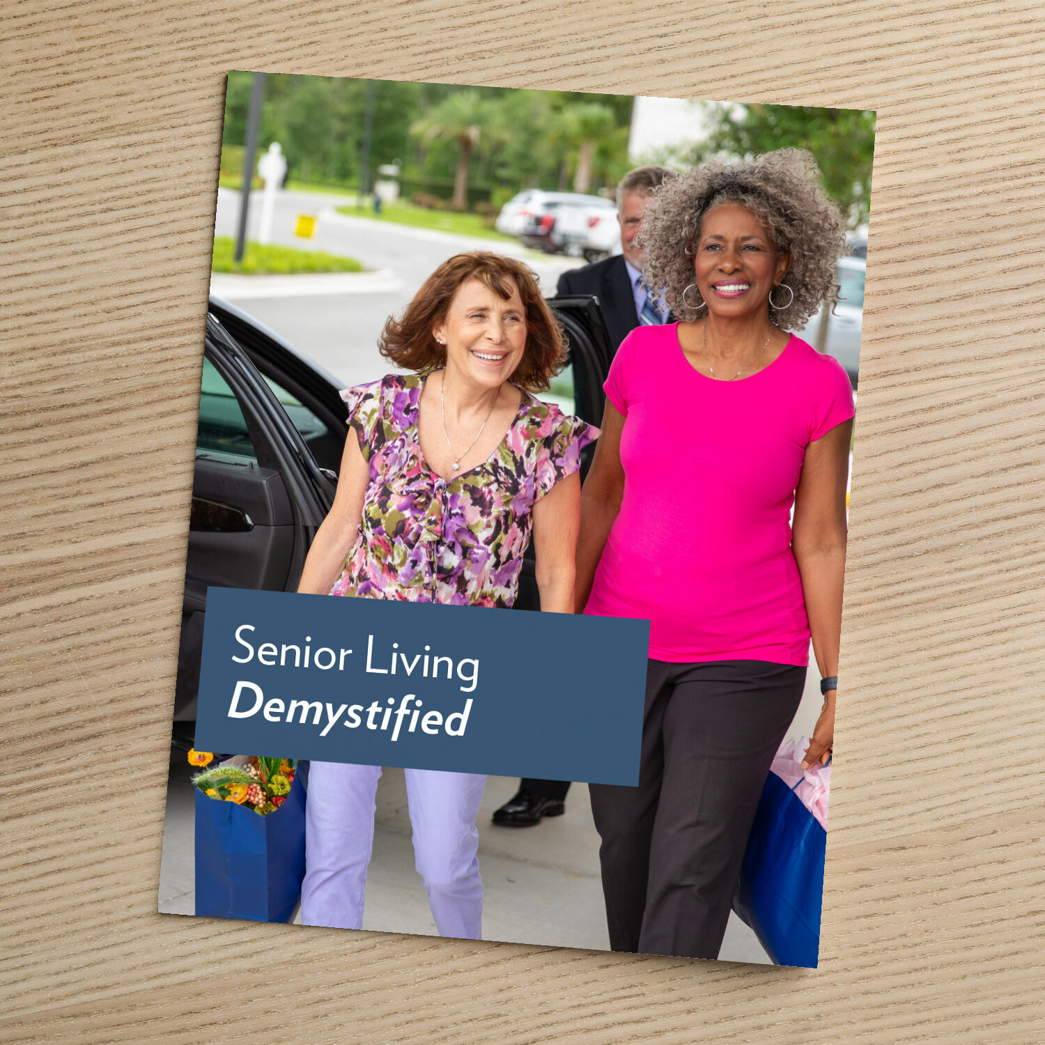 A guide titled "Senior Living Demystified" resting on a wooden surface, featuring a photo of two older women walking together and smiling outdoors.