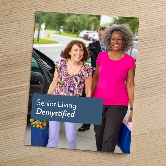 A booklet titled "Senior Living Demystified" resting on a wooden surface, featuring a photo of two older women walking together and smiling outdoors.