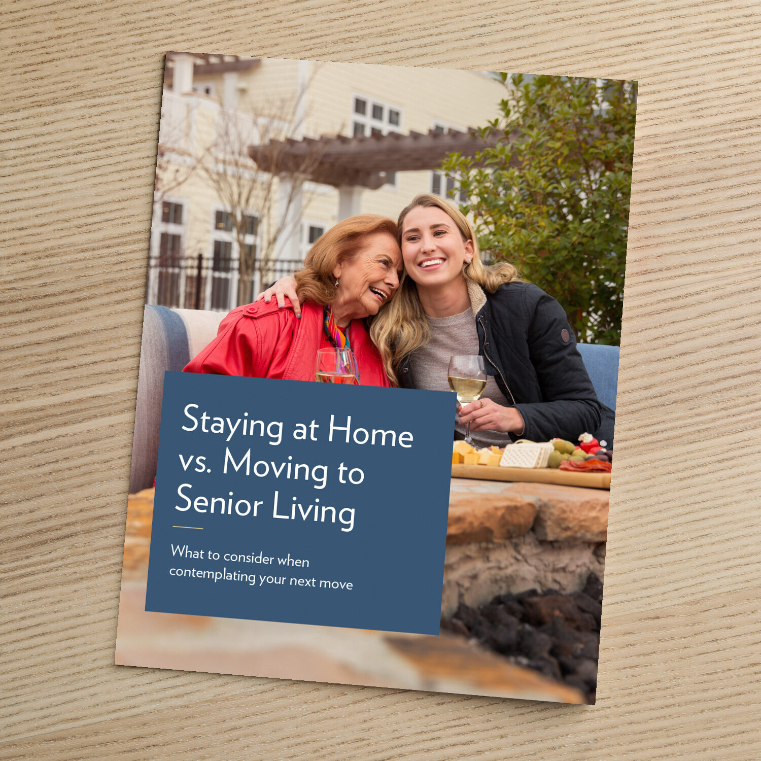 A booklet titled "Staying at Home vs. Moving to Senior Living: What to consider when contemplating your next move," resting on a wooden surface, featuring a photo of an older woman and a younger woman smiling together outdoors.
