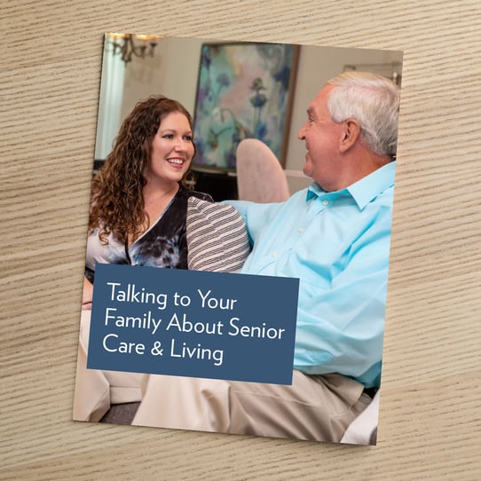 A booklet titled "Talking to Your Family About Senior Care &amp; Living" resting on a wooden surface, featuring a photo of a younger woman and an older man having a warm conversation while seated indoors.