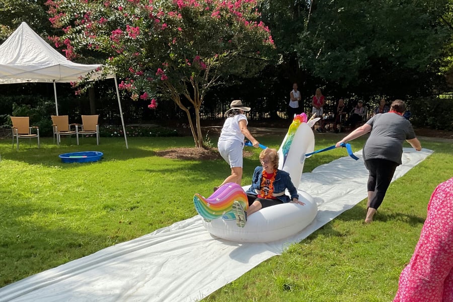 People enjoy a lively outdoor event, pulling an inflatable unicorn float across a lawn slip and slide, showcasing playful social activities at senior living in Alpharetta, GA and active senior living Alpharetta, GA.