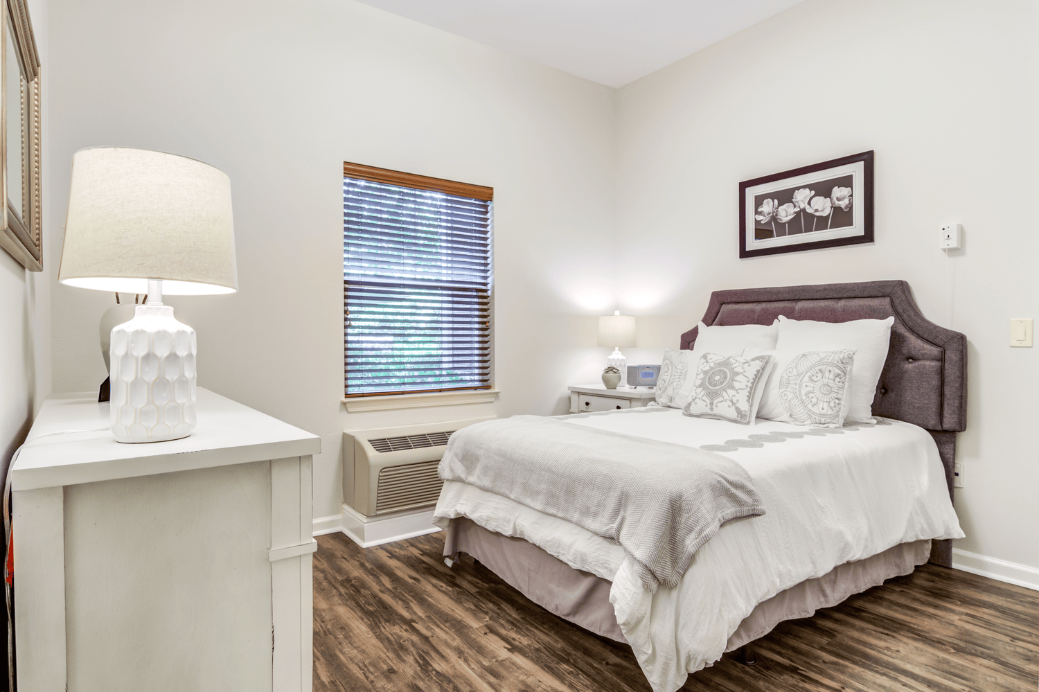 Serene model bedroom with a plush upholstered bed and soft natural light, reflecting the cozy comfort of senior living in Alpharetta, GA and senior apartments in Alpharetta, GA.