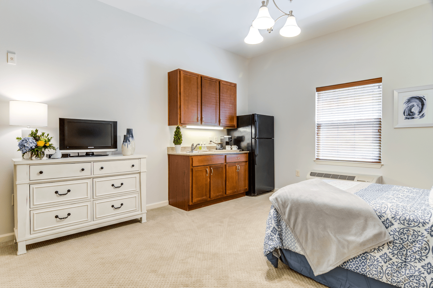 Bright model studio apartment with wood cabinetry, a cozy bed, and large windows, reflecting the comfortable living options at senior apartments in Alpharetta, GA and assisted living in Alpharetta, GA.
