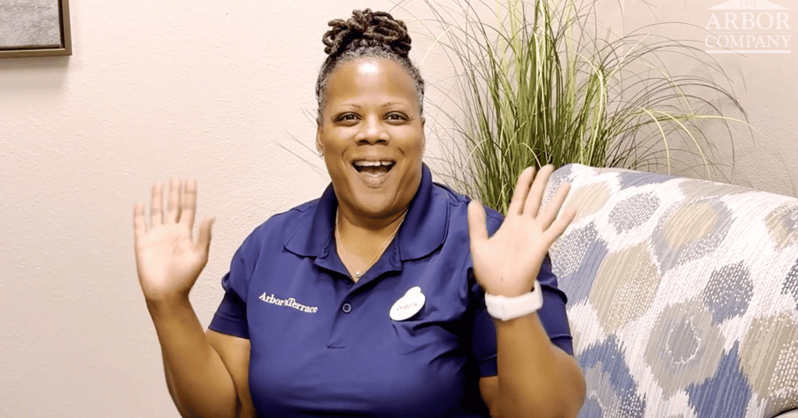 Chiquita, an Arbor Terrace team member in a navy polo and name tag, smiles broadly and waves both hands while seated on a patterned sofa beside a tall potted grass plant.