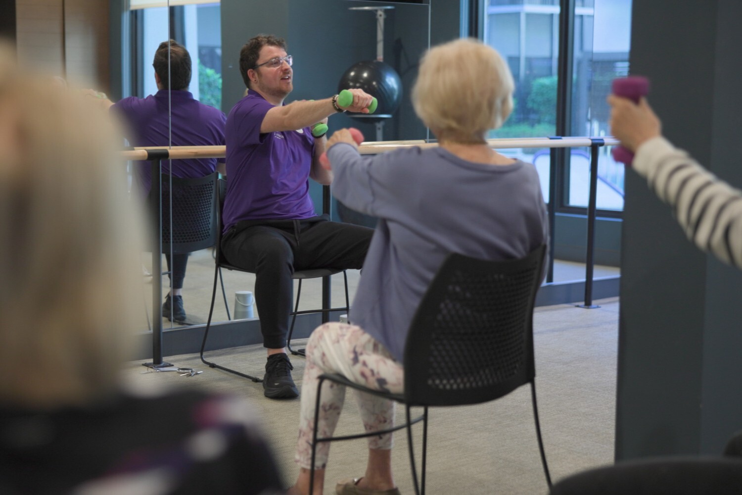 A team member in a purple shirt guides a resident through arm extensions with hand weights during a fitness class at The Arbor at Delray, showcasing the wellness programs available in this Delray Beach, FL senior living community.