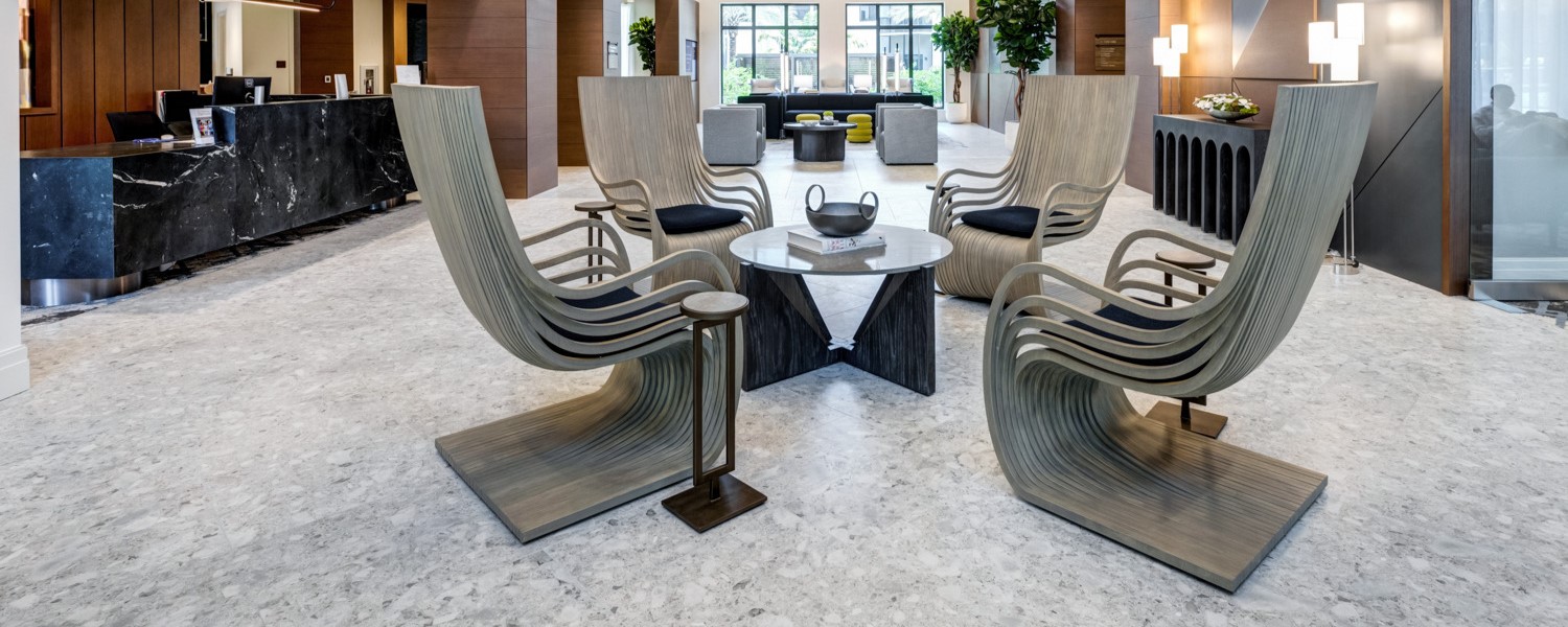 Elegant lobby seating area at The Arbor at Delray, featuring sculptural wave-back chairs arranged around a round table on marble-look flooring, with a dark stone reception desk and natural light in the background.