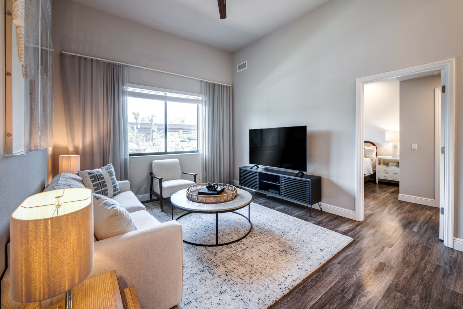 A staged living room at The Arbor at Delray features a beige sofa, round coffee table, and TV console, with a bedroom visible beyond, showcasing the comfortable senior apartments in Delray Beach, FL.
