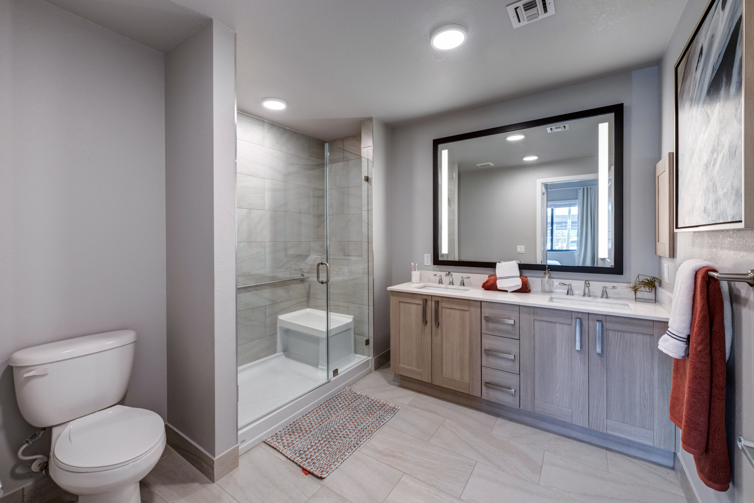 A bathroom at The Arbor at Delray features a glass-enclosed shower with a built-in bench and a dual-sink vanity with a lit mirror, showcasing the comfortable senior apartments in Delray Beach, FL.