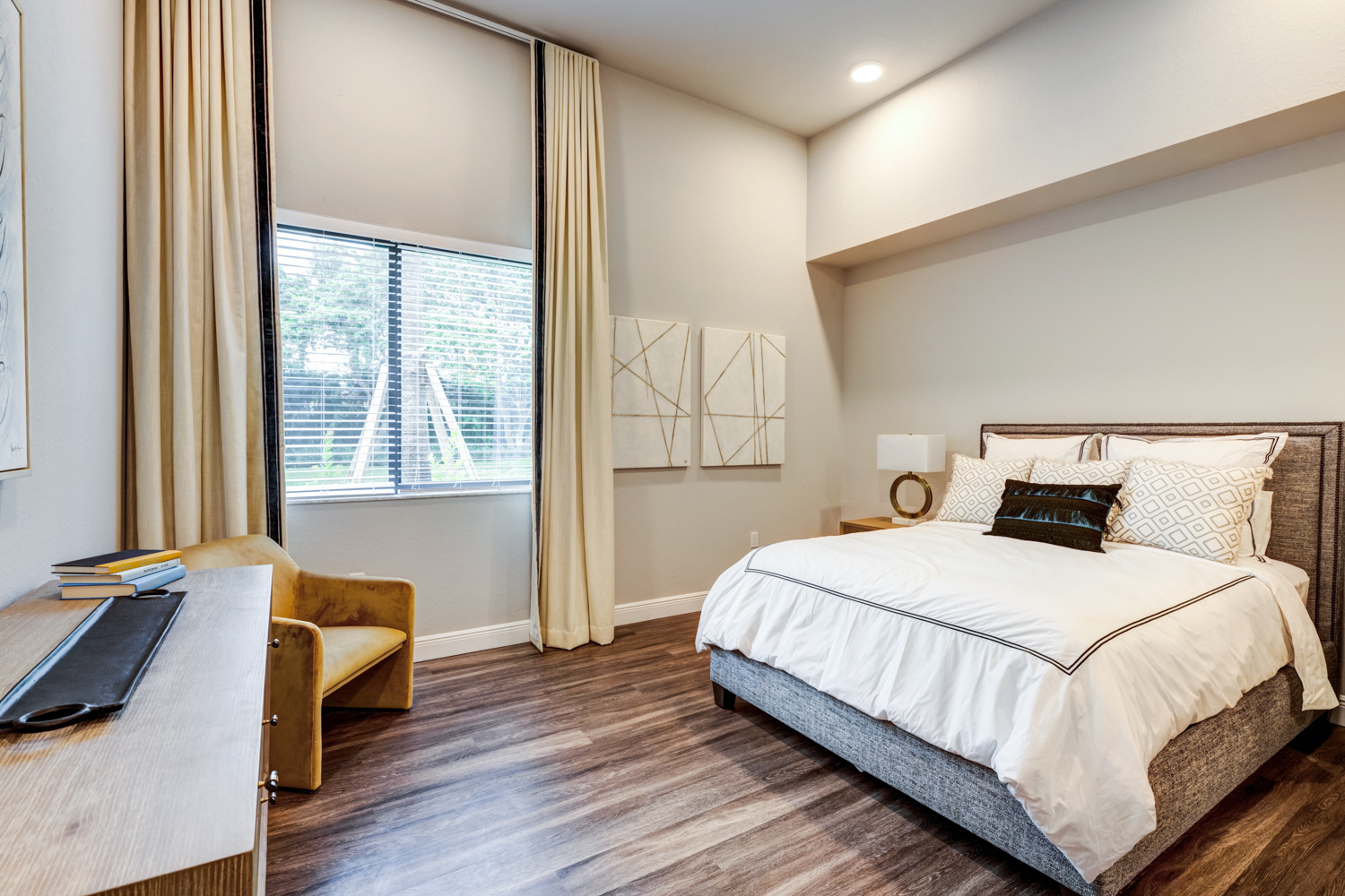 A bedroom at The Arbor at Delray features a gray upholstered headboard, cream curtains, and a mustard yellow accent chair, showcasing the comfortable senior apartments in Delray Beach, FL.