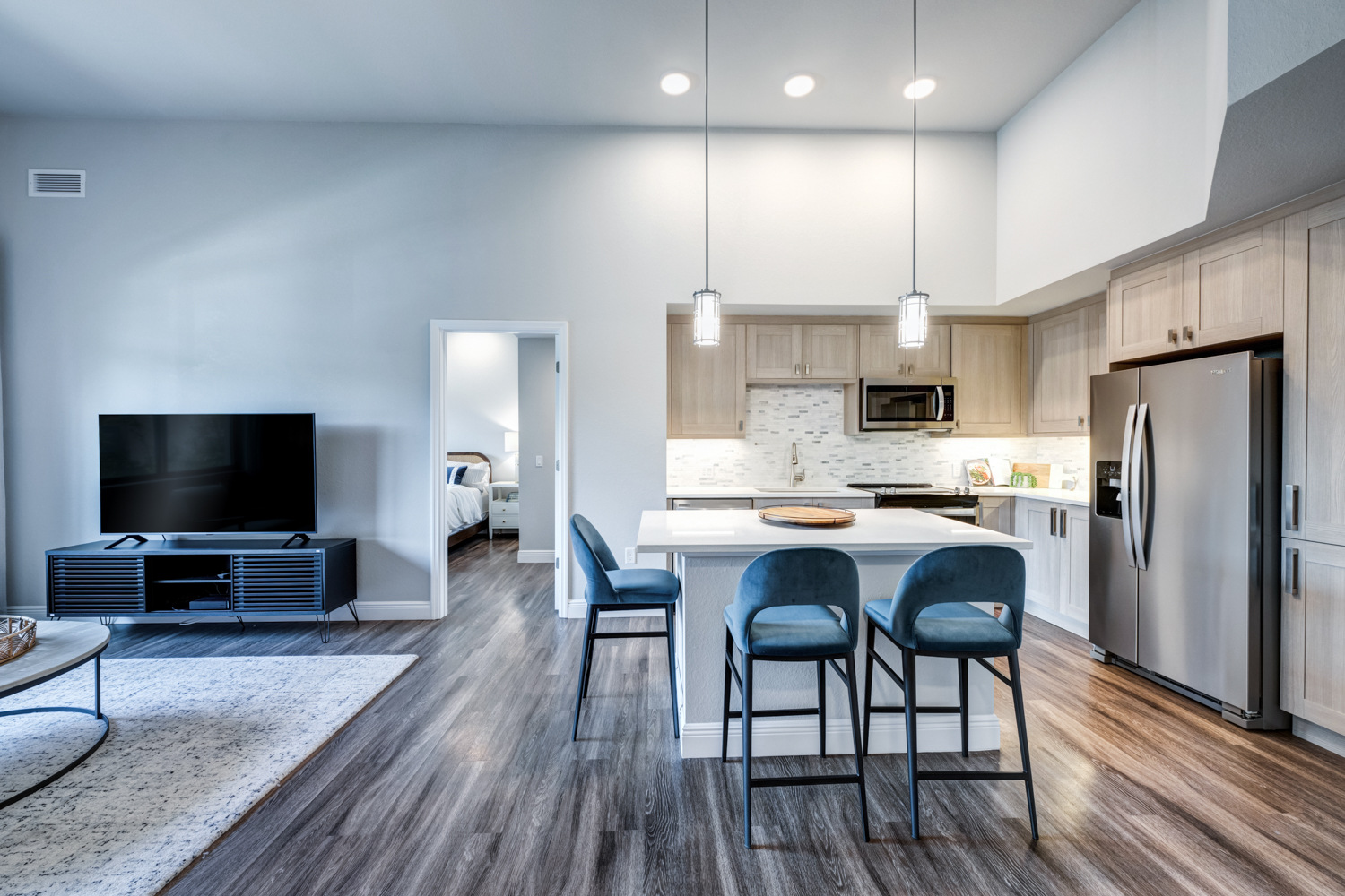 An open kitchen and living area at The Arbor at Delray features a stainless steel refrigerator, blue barstool seating at the island, and pendant lighting, showcasing the comfortable senior apartments in Delray Beach, FL.