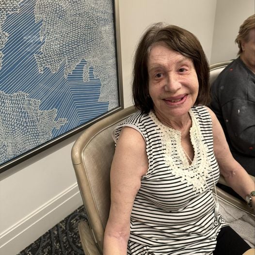 A woman smiles warmly while seated in a comfortable chair beside decorative artwork at The Arbor at Delray, reflecting the welcoming atmosphere of senior living in Delray Beach, FL.