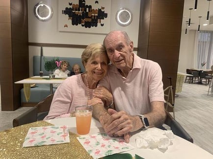 A smiling couple sits closely together at a table decorated with tropical flamingo accents, sharing a warm moment that reflects the social engagement at senior living in Delray Beach, FL.