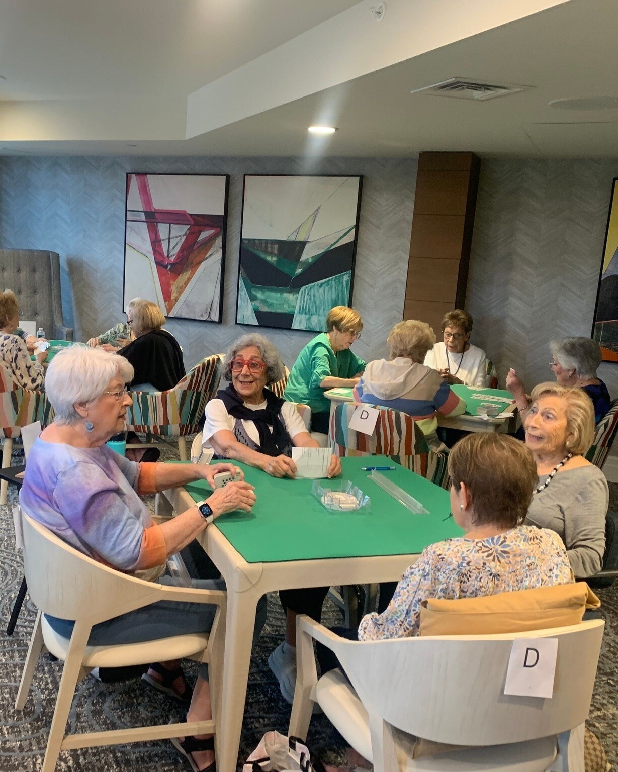 Residents gather around green-topped tables for a lively card game in a beautifully appointed common room at this senior living community in Delray Beach, FL, showcasing vibrant social engagement.