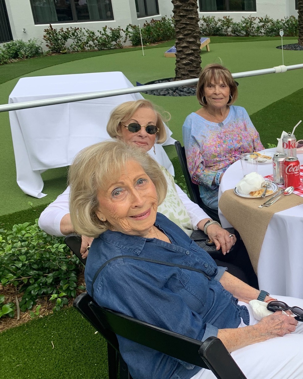 Three women enjoy time together outdoors at The Arbor at Delray, seated near the putting green and lush landscaping, reflecting the warm social engagement of senior living in Delray Beach, FL.
