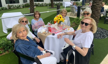 Five smiling women gather around a beautifully set outdoor table with a sunflower centerpiece, palm trees and a putting green nearby, reflecting the resort-style amenities of senior living in Delray Beach, FL.