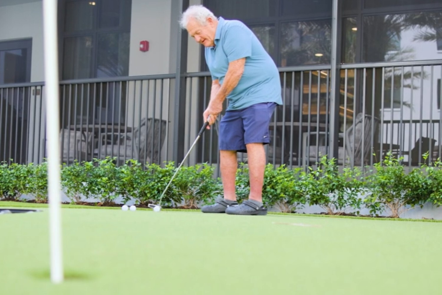 A resident enjoys a round of putting on the outdoor green at this active senior living community in Delray Beach, FL, surrounded by lush landscaping and a modern building backdrop.