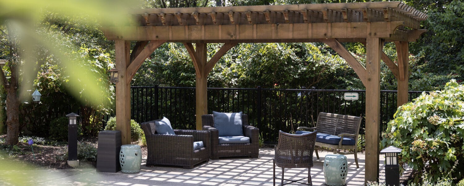 A wooden pergola over a patio seating area featuring wicker furniture with navy blue cushions, surrounded by lush greenery and black iron fencing, with decorative lanterns and ceramic garden stools accenting the space.
