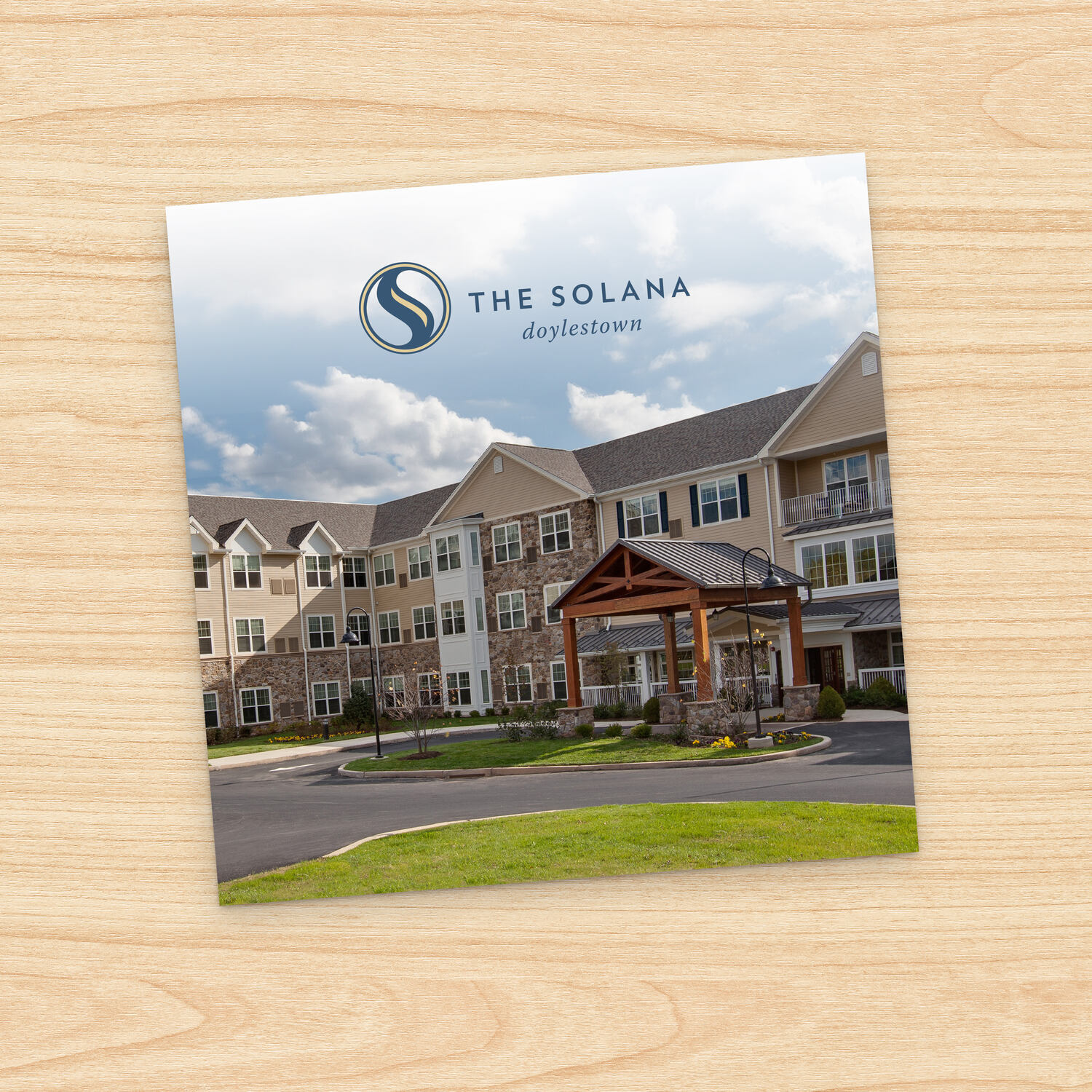 Square brochure cover for The Solana Doylestown, angled on a light wood surface. The cover photo shows a large multi-story senior living building with tan siding and stone accents, featuring a wood-beamed entrance canopy, circular driveway, and landscaped lawn under a partly cloudy sky.