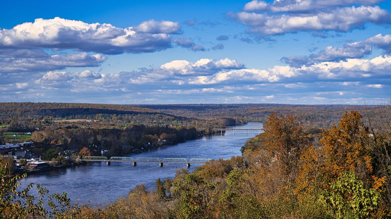 A sweeping aerial view of a wide river bordered by rolling, tree-covered hills under a bright blue sky, near senior living near the Delaware River in New Hope, PA, Bucks County.
