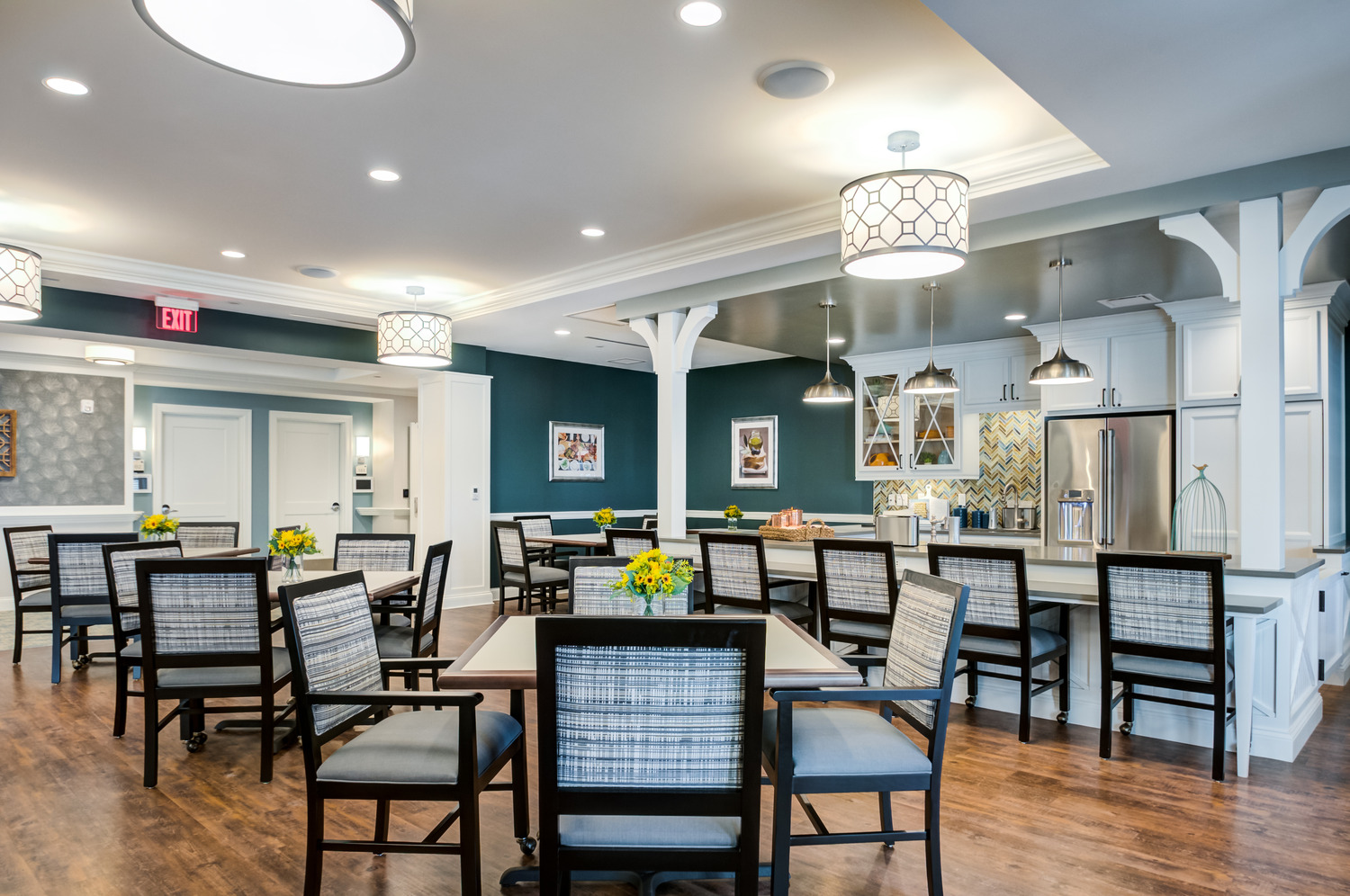 A bright dining room with a deep teal accent wall, glowing pendant lights, wood floors, and tables set with upholstered chairs and fresh yellow flowers beside an open kitchen, a welcoming space for senior living with on-site dining in Exton, PA, and assisted living near King of Prussia, PA.