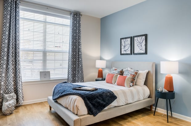 A bright apartment bedroom with a soft blue accent wall, an upholstered bed layered with a navy throw and coral pillows, coral lamps on round navy nightstands, and framed botanical prints, a cozy retreat in senior apartments in Exton, PA, and assisted living in Phoenixville, PA.