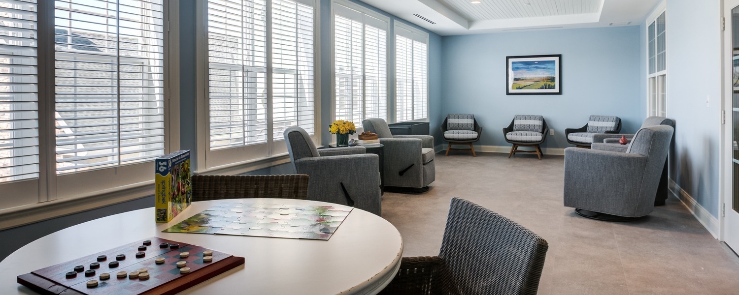 A sunlit game room with walls of white shuttered windows, soft blue walls, gray upholstered armchairs, and a round table set with board games and woven chairs, a relaxing gathering space for personal care in Exton, PA, and assisted living near Chester Springs, PA.