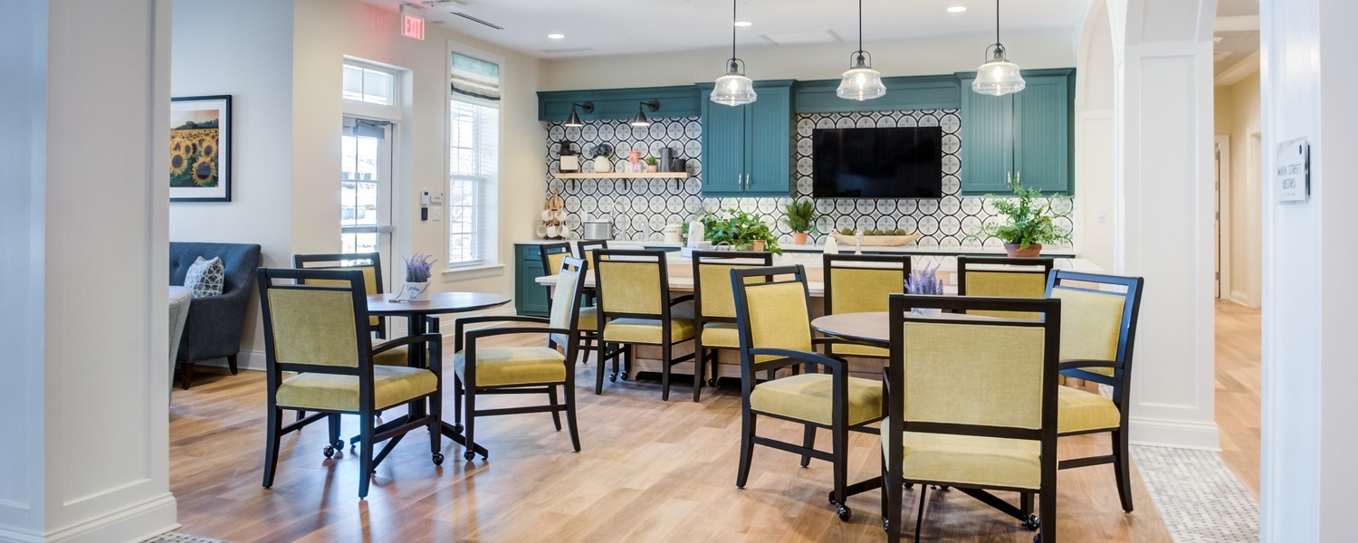 A bright dining area with teal cabinetry, a patterned tile backsplash, glowing pendant lights, and round tables with yellow upholstered chairs on warm wood floors, a cheerful spot to gather at personal care communities near Paoli, PA, and assisted living in Malvern, PA.
