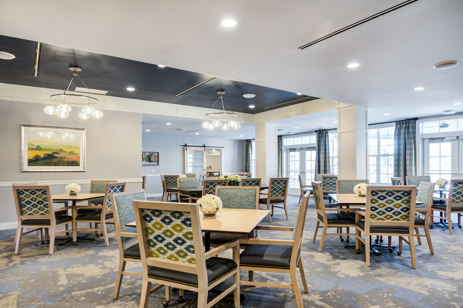 A spacious dining room with square tables, patterned blue and green chairs, white hydrangea centerpieces, glass globe chandeliers, and French doors filling the space with light, a welcoming place to dine at assisted living communities in Exton, PA, and assisted living near West Chester, PA.