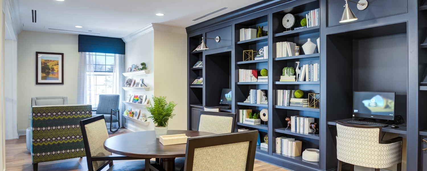 A bright library lounge for senior living in Exton, PA, with navy built-in bookshelves, two computer stations, a round table with a potted plant and woven chairs, and a cozy seating nook by the window, a welcoming spot for assisted living in Exton, PA.