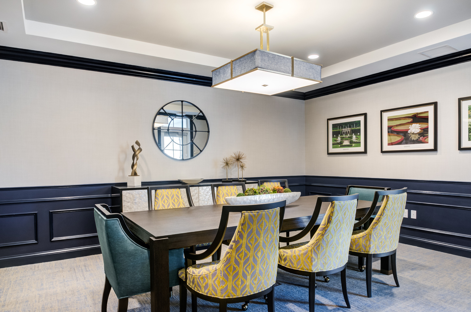A private dining room with a long dark wood table, yellow patterned and teal upholstered chairs, a square pendant light, navy wainscoting, and framed garden prints, an elegant space for gatherings at resort-style senior living Exton, PA, and assisted living in Downingtown, PA.