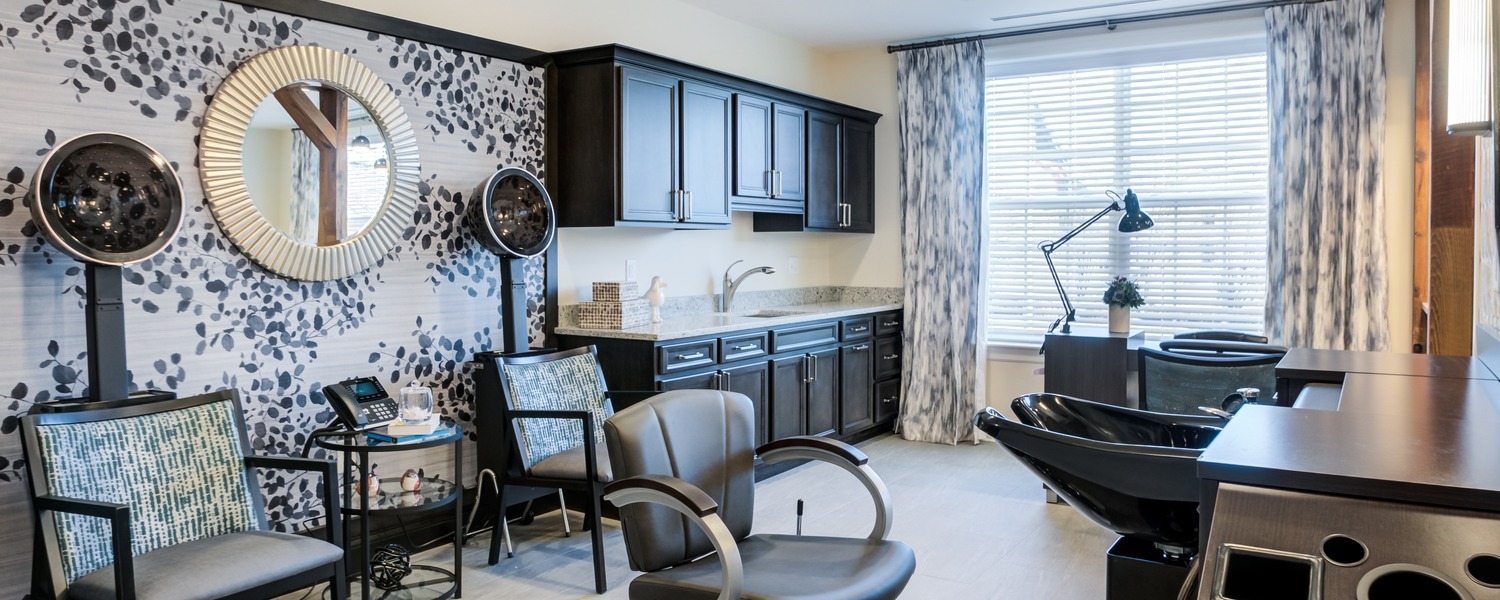 A stylish salon with a styling chair, hooded dryers, a shampoo station, and a manicure table, set against bold patterned wallpaper, a round mirror, and bright draped windows, a pampering space for wellness-focused senior living North Shore and assisted living Highland Park IL.