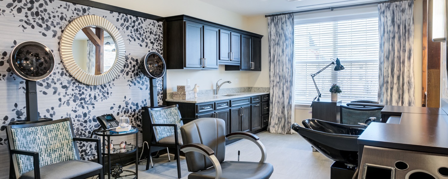 A stylish salon with hooded hair dryers, a sunburst mirror on bold leaf-print wallpaper, a styling chair, a shampoo station, and dark cabinetry by a bright window, a pampering space for assisted living in Downingtown, PA, and assisted living in Paoli, PA.
