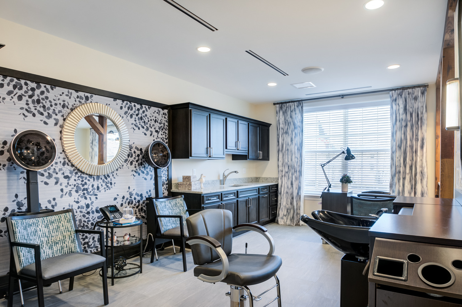 A stylish salon with hooded hair dryers, a sunburst mirror on bold leaf-print wallpaper, a styling chair, a shampoo station, and dark cabinetry by a bright window, a pampering space for assisted living in Downingtown, PA, and assisted living in Paoli, PA.