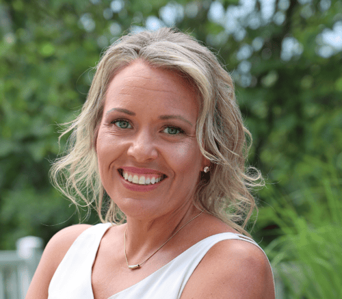 Heather Goodno, Director of Sales, smiles brightly with wavy blonde hair and a white top against a leafy green backdrop, helping families explore senior living near West Chester, PA, and personal care in Exton, PA.
