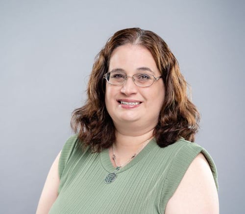 Bernadette Kehlmann, Business Office Director, smiles in glasses and a green top against a soft gray backdrop, supporting families at senior living near Chester Springs, PA, and senior living in Downingtown, PA.