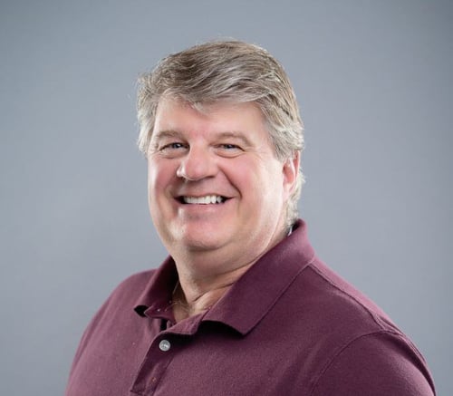Chuck Niccolls, Engagement Director, smiles in a maroon polo against a soft gray backdrop, bringing energy to active senior living near Downingtown, PA, and senior living near Phoenixville, PA.