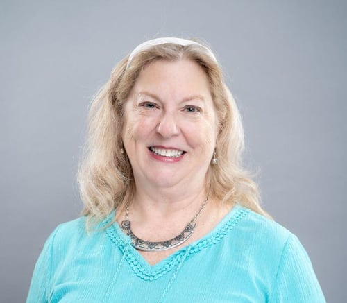 Donna Greiss, Resident Care Director, smiles in a turquoise top and silver necklace against a soft gray backdrop, supporting personal care with daily assistance in Exton, PA, and senior living near Paoli, PA.