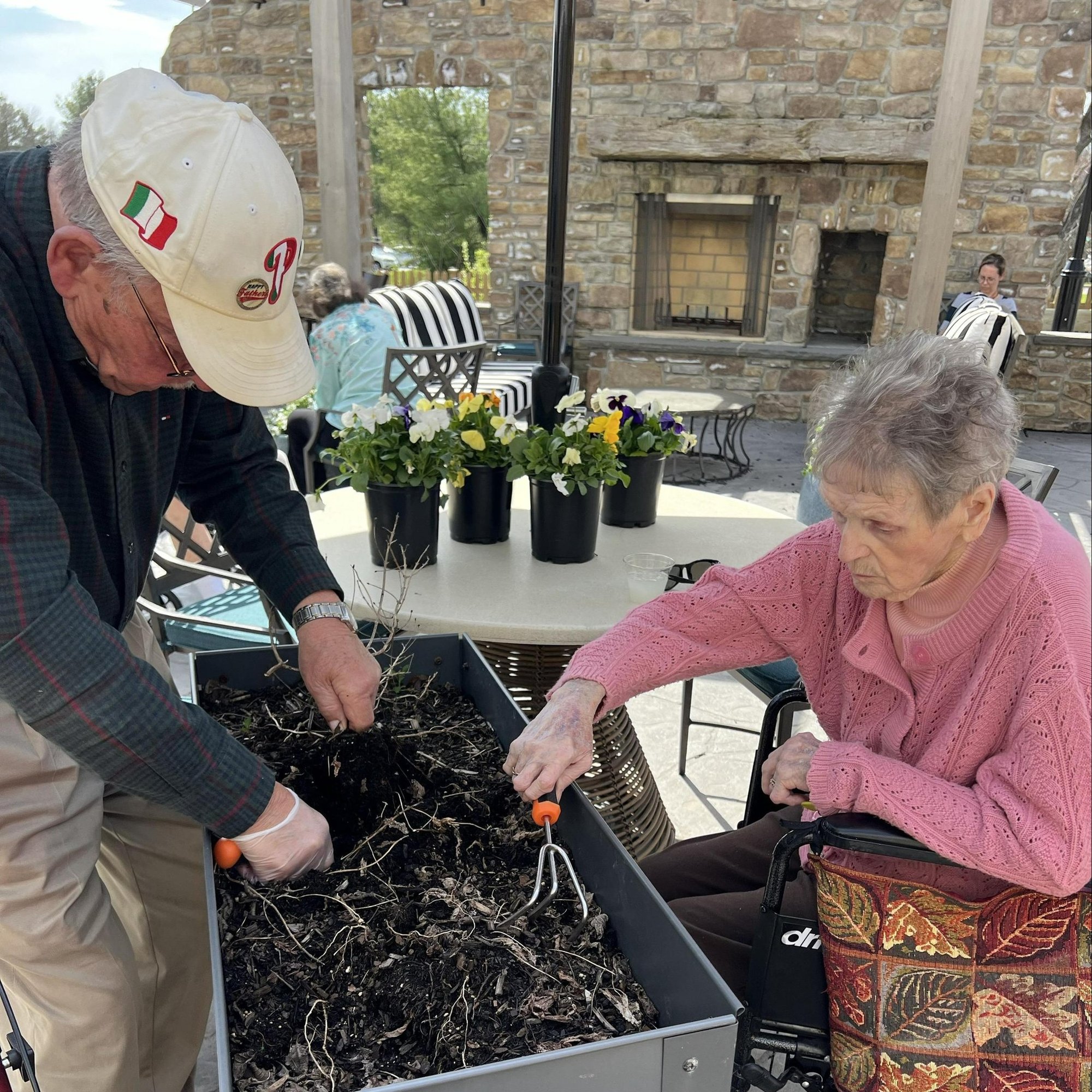 A man in a ball cap and a woman in a pink sweater tend a raised garden bed on a sunny stone patio lined with potted flowers, enjoying hands-on gardening at senior living near Chester Springs, PA, an inviting option for those exploring senior living in Malvern, PA.