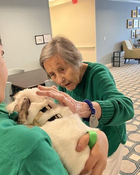 A woman in a green sweater gently pets a fluffy white dog held by the person beside her in a bright common area, a sweet moment of connection at pet-friendly senior living in Exton, PA, and a caring community for those seeking senior living in Downingtown, PA.