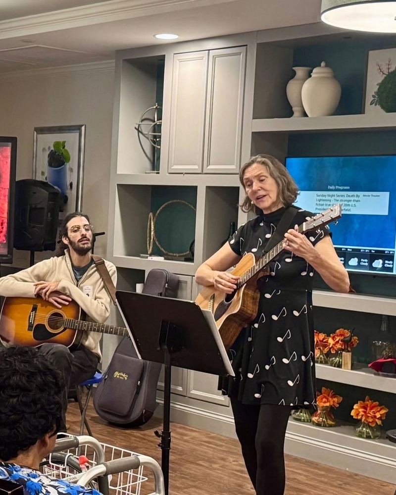 Two musicians play acoustic guitars during a lively performance, one standing in a polka dot dress and one seated beside built-in shelves, bringing warm live music to senior living in Exton, PA, a welcoming choice for senior living near West Chester, PA.