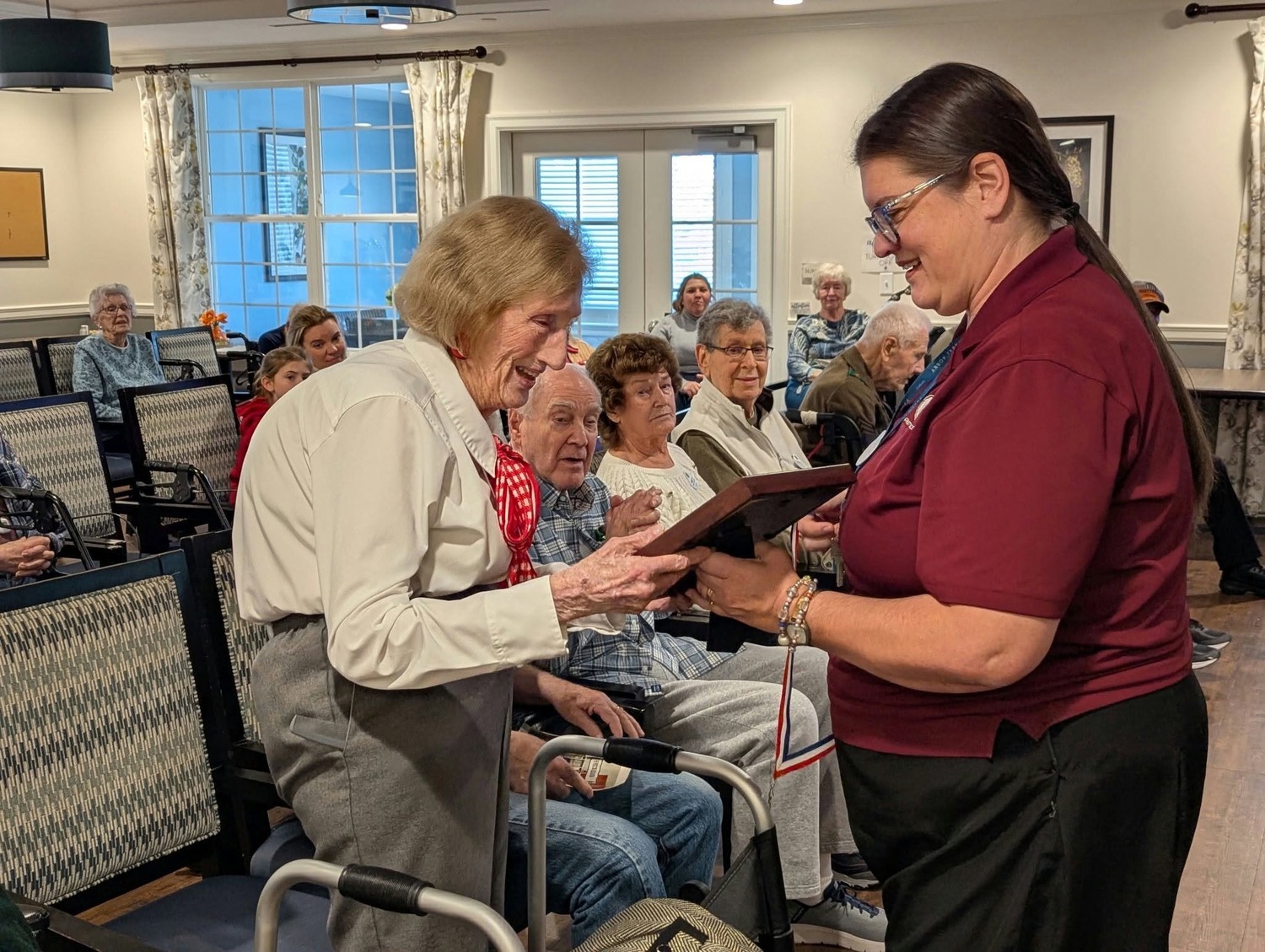 A resident proudly receives a special award from a smiling staff member in a lively community room filled with fellow residents, a proud milestone within senior living near South Coventry, PA and senior living in Exton, PA.