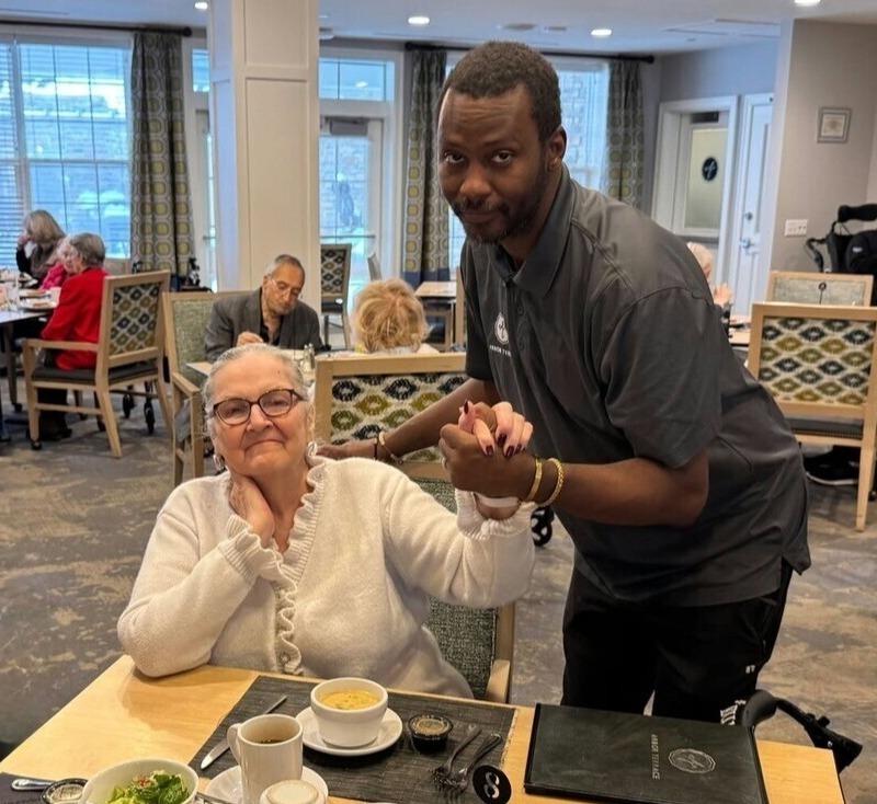 A smiling woman in a white cardigan holds hands with a man in a dark shirt who leans in beside her at a dining table, sharing a warm moment in a bright dining room at senior living with on-site dining in Exton, PA, a friendly place for senior living near Paoli, PA.